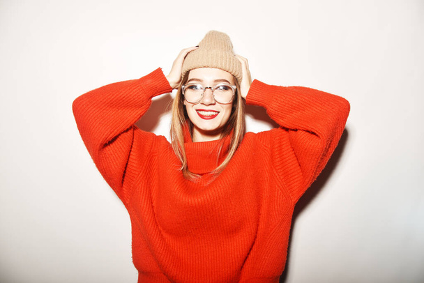 A girl in a red sweater and hat, a beauty with glasses for sight, having fun, dancing, a hipster girl is bright. Model with a caret smiles and laughs in the studio on a white background. - Foto, imagen