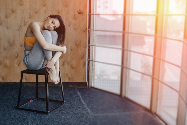 Νεαρό ελκυστικό κορίτσι γυμναστικής κάθεται στην καρέκλα κοντά στο παράθυρο στο φόντο ενός ξύλινου τοίχου, στηρίζεται σε μαθήματα γιόγκα στο γυμναστήριο - Φωτογραφία, εικόνα