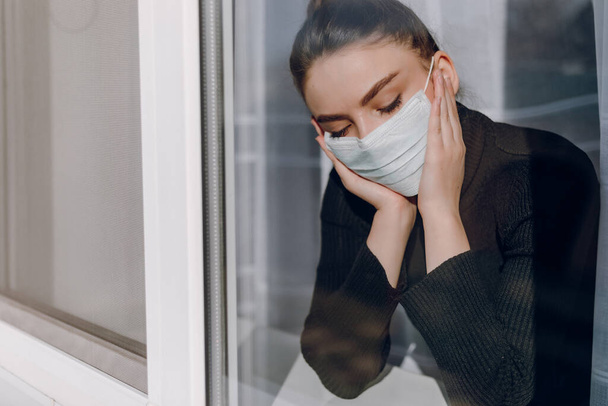jong aantrekkelijk meisje met een beschermend medisch masker kijkt uit het raam. isolatie tijdens de epidemie. isolatie van medische tehuizen. - Foto, afbeelding