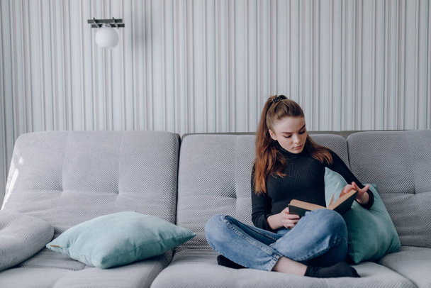 young attractive girl on the couch reads a paper book. mental development. useful use of time at home. warm home comfort. - Φωτογραφία, εικόνα