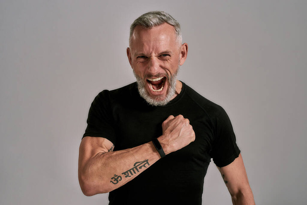 Middle aged muscular man in black t shirt shouting at camera, beating his chest while posing in studio over grey background - Fotoğraf, Görsel