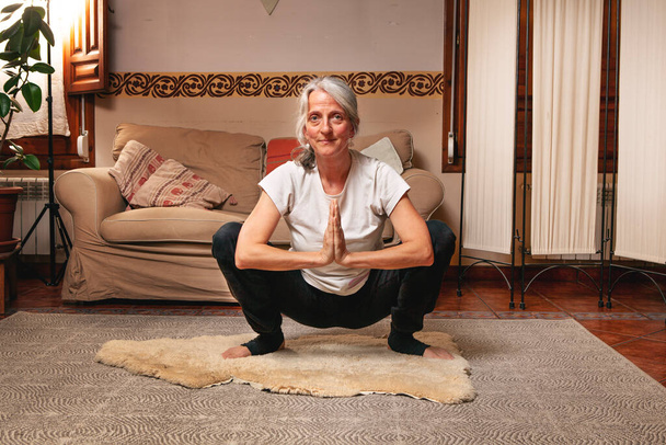 vrouw van middelbare leeftijd met grijs haar die kundalini yoga beoefent in haar woonkamer - Foto, afbeelding