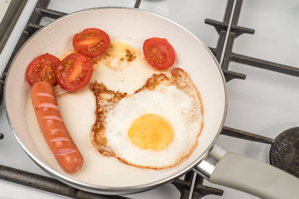 Petit déjeuner facile et rapide - un œuf, des tomates et des saucisses sont frits dans une casserole avec un revêtement antiadhésif en céramique blanche - Photo, image