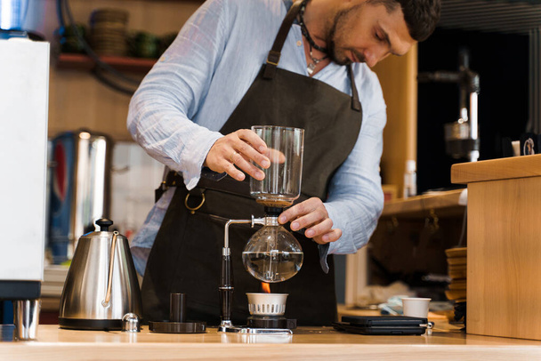 Όμορφος γενειοφόρος barista κατέχει Syphon συσκευή στα χέρια πριν από την παρασκευή καφέ στο καφέ. Syphon εναλλακτική μέθοδος παρασκευής καφέ. Σκανδιναβική μέθοδος παρασκευής καφέ. - Φωτογραφία, εικόνα