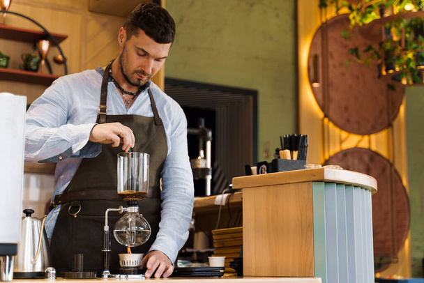 Handsome bearded barista mix freshly ground coffee in syphon device for coffee brewing in cafe. Syphon alternative method of making coffee. Scandinavian method of coffee making. - Foto, Imagem