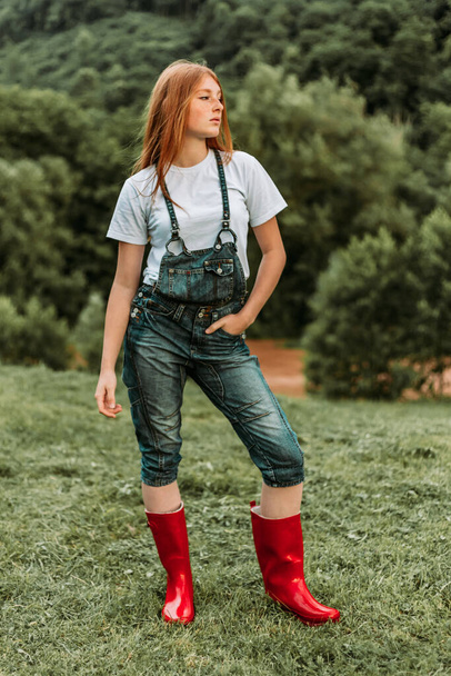 Photo of ginger girl in jeans with suspenders and red boots holding hand in her pocket - Photo, image