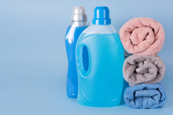 Flaschen Waschmittel und Weichspüler mit sauberen Handtüchern auf blauem Hintergrund. Behälter mit Reinigungsmitteln, Haushaltschemikalien. Flüssigwaschmittel und Conditioner. Wäschetag, Reinigungskonzept - Foto, Bild
