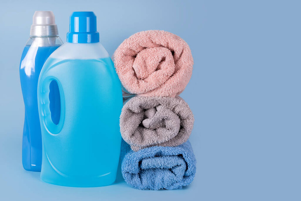 Garrafas de detergente e amaciador de tecido com toalhas limpas no fundo azul. Recipientes de produtos de limpeza, produtos químicos domésticos. Detergente líquido e condicionador. Dia de lavanderia, conceito de limpeza
 - Foto, Imagem