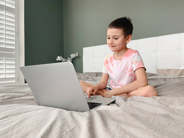 Běloch chlapec dítě sedí v posteli a učí se on-line na notebooku internetu. Virtuální lekce na videu. Samostatná izolace doma. Vzdálená vzdálená výuka videa. Nová normální školní studie pro děti. - Fotografie, Obrázek