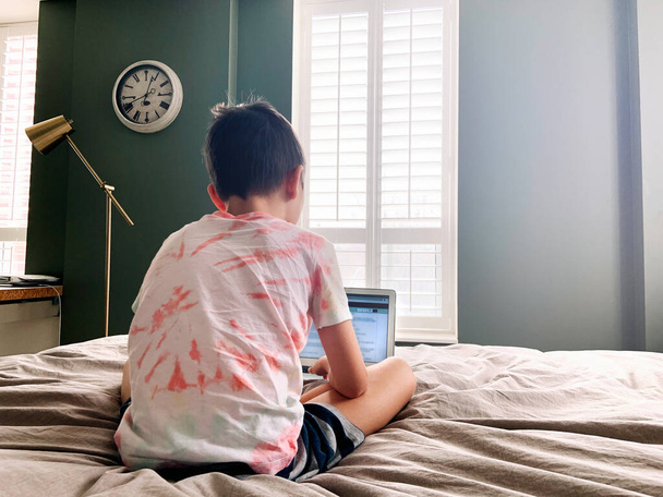 Kaukasische jongen kind zitten in bed en leren online op laptop Internet. Virtuele les op video. Zelfisolatie thuis. Afstandsonderwijs op afstand. Nieuwe normale school studie voor kinderen. - Foto, afbeelding