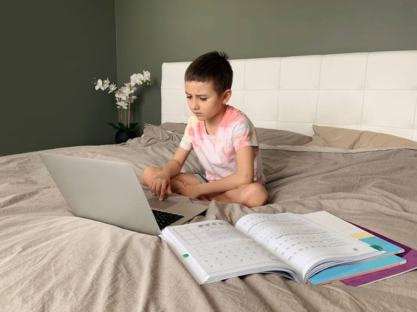 Καυκάσιο παιδί που κάθεται στο κρεβάτι και μαθαίνει στο διαδίκτυο στο φορητό υπολογιστή. Μάθημα εικονικής τάξης σε βίντεο. Αυτοαπομόνωση στο σπίτι. Απομακρυσμένη εκπαίδευση βίντεο. Νέα κανονική σχολική μελέτη για παιδιά. - Φωτογραφία, εικόνα