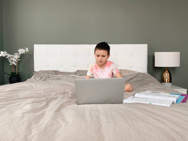 Kaukázusi fiú gyermek ül az ágyban, és tanul online laptop interneten. Virtuális óra videón. Önzárkózottság otthon. Távoli videó oktatás. Új normál iskolai tanulmány gyerekeknek. - Fotó, kép