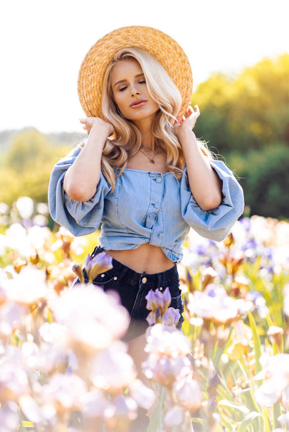Beautiful blonde woman with wavy long hair wearing summer hat and posing near the flowers - Valokuva, kuva