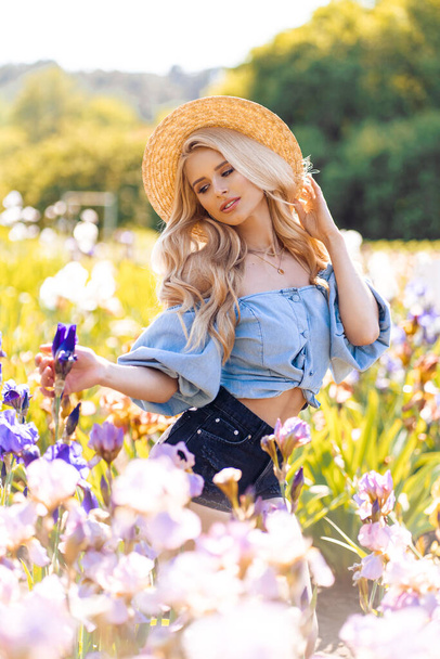 Piękna blondynka z falowanymi długimi włosami, nosząca letni kapelusz i pozująca w pobliżu kwiatów - Zdjęcie, obraz