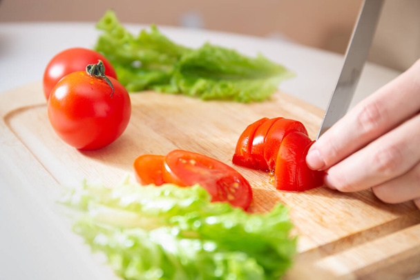 Las manos femeninas cortan un jugoso tomate rojo en rodajas con un cuchillo en una tabla de cortar de madera. Un método de preparación de verduras e ingredientes antes de cocinar
. - Foto, Imagen