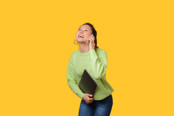 Feliz mujer sonriente está hablando por teléfono mientras sostiene su computadora portátil sobre el fondo amarillo
 - Foto, imagen