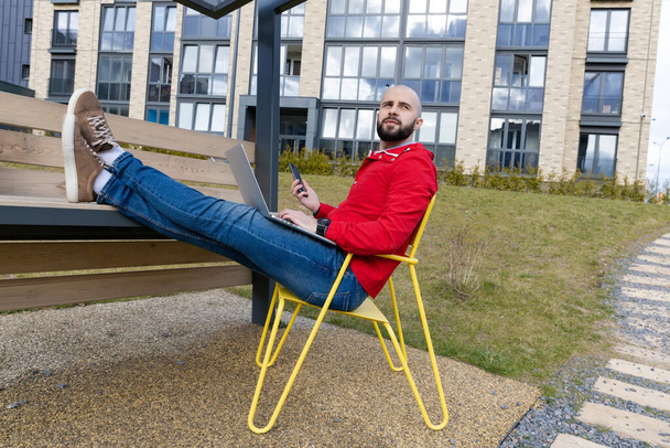 A brutal guy with a beard in casual clothes works at a computer in a residential quarter. Concept of working outside the home - Foto, afbeelding