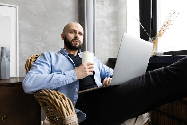 Un tipo con una camisa azul está trabajando en un portátil en casa junto a la ventana y bebiendo de un vaso. El concepto de trabajar a distancia - Foto, imagen