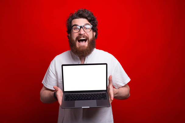 Hipster barbu excité montre écran vide d'un ordinateur portable sur fond rouge. - Photo, image