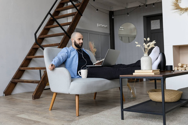 Chlápek v modré košili a černých džínách sedí na gauči a pracuje u počítače. Koncept práce na volné noze - Fotografie, Obrázek