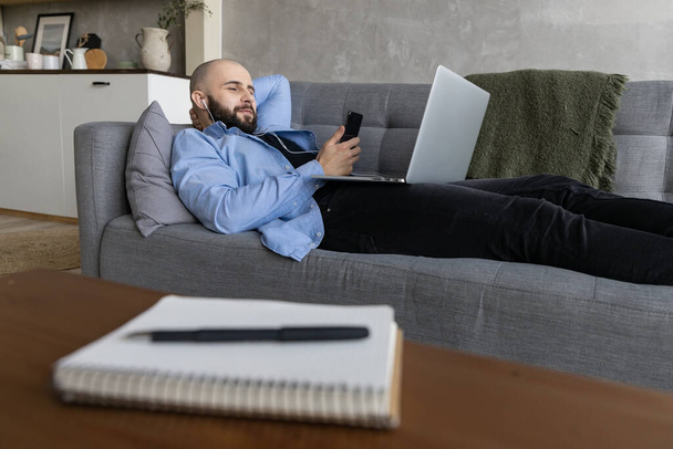 Парень лежит на диване, работает над ноутбуком и разговаривает по телефону. Концепция работы из дома - Фото, изображение