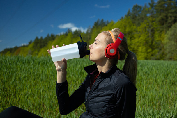 A girl is sitting on a fitness mat listening to music and drinking water after a workout. Against the background of a green field and forest - Photo, Image