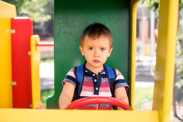 Future driver: toddler boy with serious face playing as truck driver at outdoor playground. Little kid with backpack sitting at steering wheel and driving toy vehicle in park. Child dreams concept - Foto, Imagen