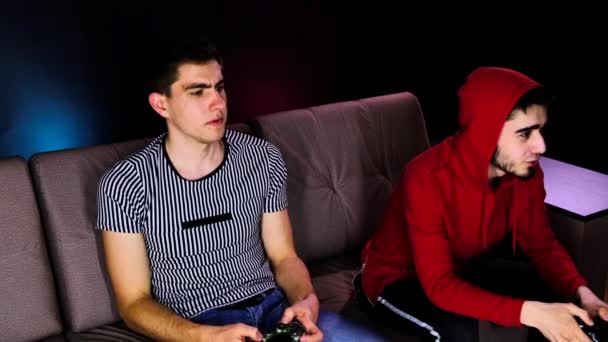 Emotional two young guys are sitting on the couch in front of the TV and excited playing a game console, holding a joystick. Mens gaming with a wireless controller. Cozy room is lit with neon light. - Felvétel, videó