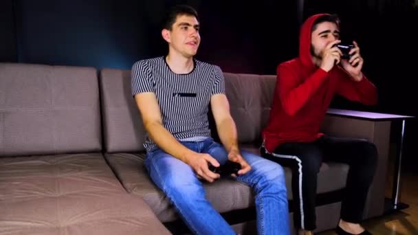 Having fun enjoy victory. Emotional two young guys are sitting on the couch in front of the TV and excited playing a game console, holding a joystick. Mens gaming with a wireless controller. Cozy room - Filmmaterial, Video