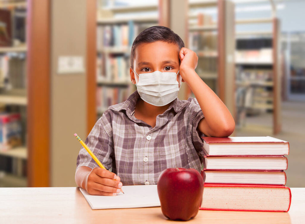 Niño hispano usando máscara facial con libros, manzana, lápiz y papel en la biblioteca
. - Foto, imagen