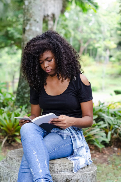 eine junge schwarze Frau zwischen 20 und 30 Jahren, die allein in einem Park sitzt und ein Buch liest - Foto, Bild