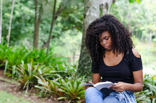 eine junge schwarze Frau zwischen 20 und 30 Jahren, die allein in einem Park sitzt und ein Buch liest - Foto, Bild