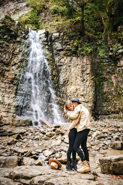 Young and beautiful couple at the mountain waterfall - Happy tourists visiting mountains. Lovestory. Tourists in hats. Military fashion - Фото, зображення