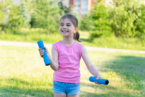 The child goes in for sports in the park. Little girl is engaged with dumbbells. - Фото, зображення