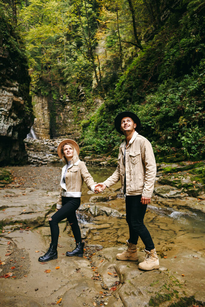 Young and beautiful couple at the mountain waterfall - Happy tourists visiting mountains. Lovestory. Tourists in hats. Military fashion - Photo, Image