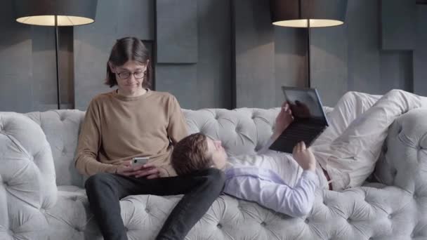 Muž v brýlích píše na smartphone jako jeho partner pomocí notebooku. Portrét mladého kavkazského homosexuálního páru odpočívajícího doma. Koncept stejného sexuálního vztahu, závislost na zařízení, životní styl lgbt. - Záběry, video