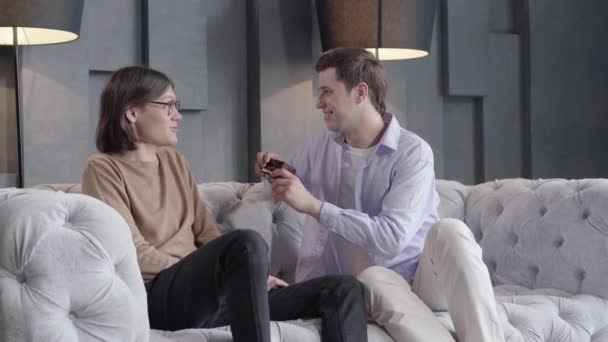 Smiling happy man giving present to gay partner on anniversary. Portrait of handsome young same sex Caucasian couple celebrating holiday at home. Lgbt, love, romance, happiness. - Séquence, vidéo