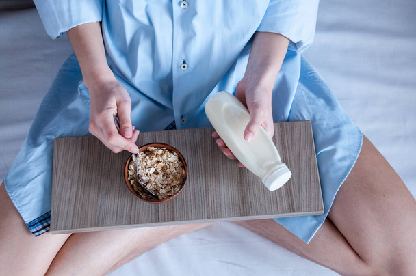 Breakfast in bed, a girl in a blue shirt sitting on a white sheet and Breakfast cereal with milk. Girl holding a bottle of milk. - Φωτογραφία, εικόνα