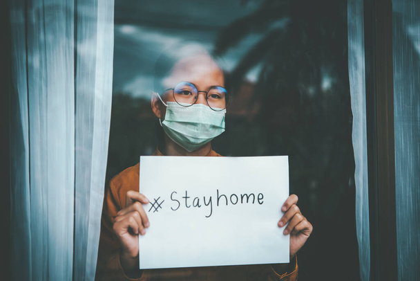 Azjatka nosząca okulary, nosząca maskę, trzymająca papier o nazwie # stay home, jest znudzona koniecznością samodzielnego zatrzymywania i leczenia choroby w domu. Koncepcja kwarantanny domowej, zapobieganie COVID-19 - Zdjęcie, obraz