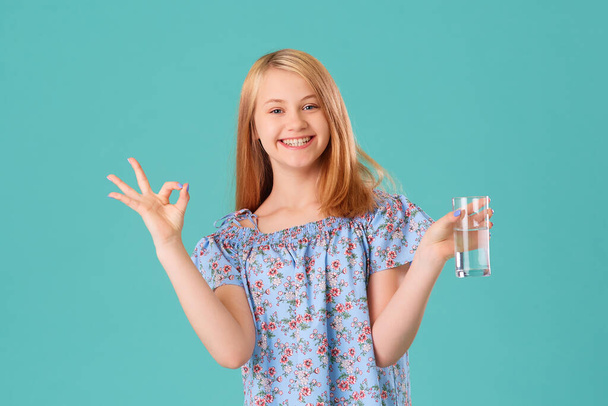 Happy smiling teen girl with a glass of water, she adheres to a drinking regime. Studio portrait on a turquoise background. Concept of health care and replenishment of the water-salt balance - Foto, Bild