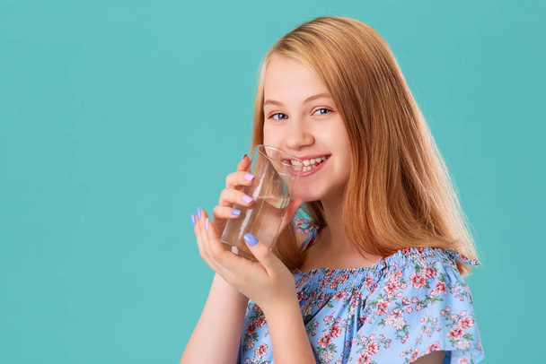 Portrait of a smiling girl drinking water from a glass glass. On a turquoise background - Foto, Bild