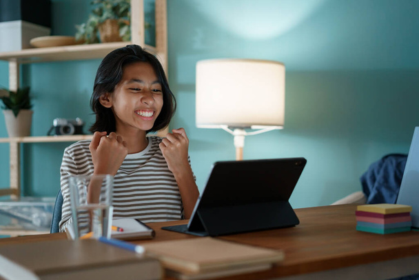 Asian little girl successfully is studying online via the internet on a digital tablet while sitting on a desk at home night. Asia child happy and smile. Concept of online learning at home - Foto, Imagem