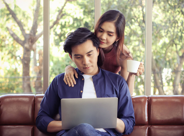 Young Asian couple in living room , a man sitting on couch using computer on his lap while woman standing behind ,holding coffee cup and looking at computer screen together.  - Φωτογραφία, εικόνα
