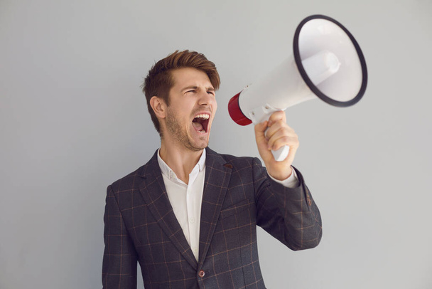 Stylish executive man screaming with loudspeaker on gray background - 写真・画像