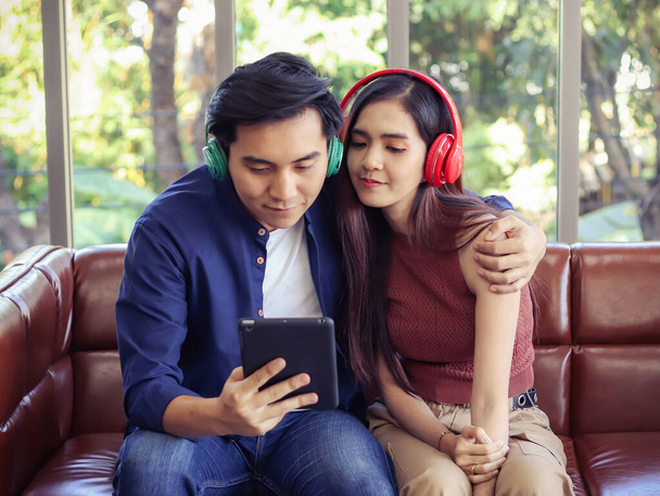 jong en aantrekkelijk aziatisch paar dragen koptelefoon zitten dicht bij elkaar op de bank in woonkamer thuis, man knuffelen vrouw, luisteren naar muziek en kijken naar tablet. - Foto, afbeelding