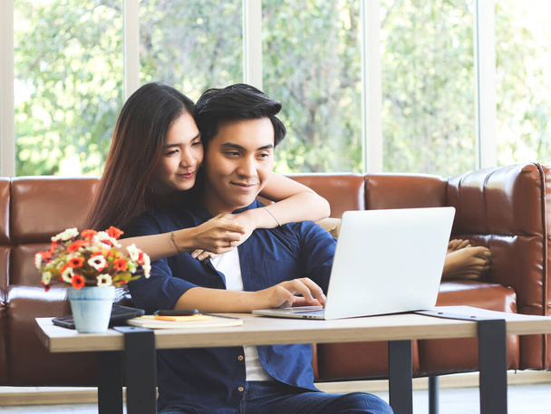 Young Asian couple spend good time at home , a man sitting on the floor with computer laptop on table while his girlfriend lying on sofa behind.work from home concept. - Фото, изображение