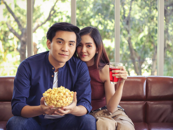 Joven y atractiva pareja asiática enamorada sentados juntos en el sofá en la sala de estar un hombre sosteniendo palomitas de maíz y una mujer sosteniendo la bebida roja
. - Foto, imagen