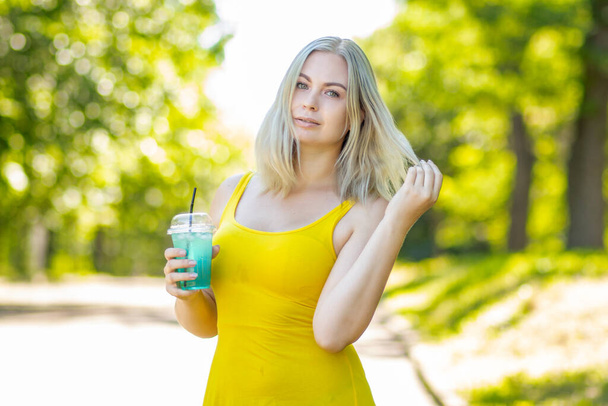 attractive stylish blond smiling woman in yellow short dress, summer fashion outfit, drinking cocktail in park - Fotografie, Obrázek