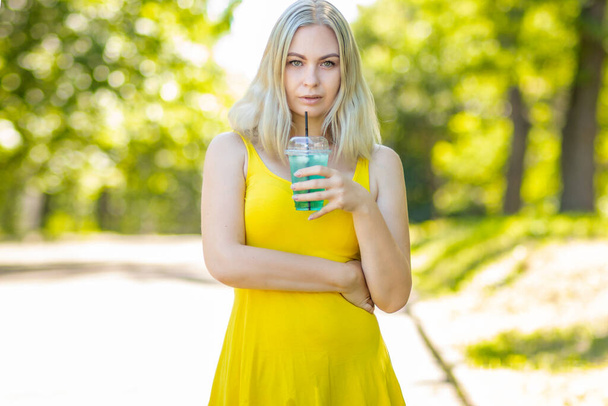 attractive stylish blond smiling woman in yellow short dress, summer fashion outfit, drinking cocktail in park - Photo, image