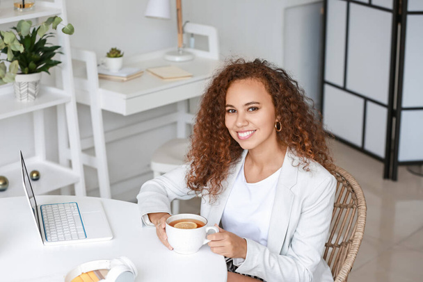 Kaunis nuori afroamerikkalainen nainen kannettavan tietokoneen kanssa juo kuumaa teetä kotona - Valokuva, kuva